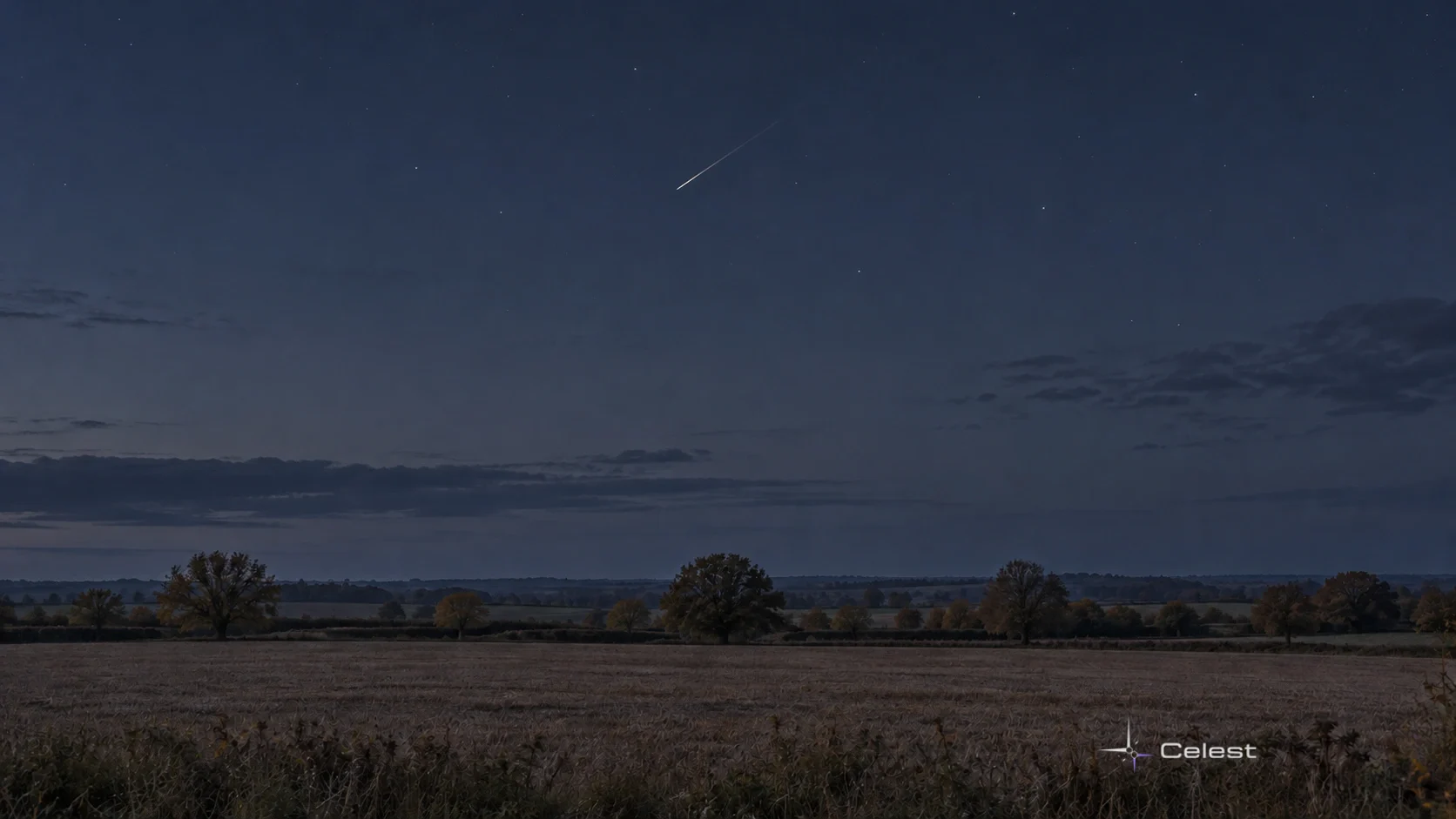 Un meteor subțire și o urmă luminoasă discretă deasupra unui câmp de toamnă, sub un cer liniștit.